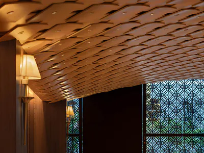 Interior view of a sophisticated Japanese-style restaurant featuring a long upholstered banquette, round wooden pedestal tables, and matching armchairs with cream upholstery, all beneath an elaborate sculptural wooden ceiling with integrated lighting and decorative lattice window screens in the background.