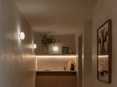 A narrow minimalist hallway with textured beige walls, a floating wooden console under a backlit shelf, spherical wall sconces, and a framed abstract artwork on the right.