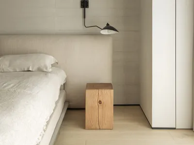 Modern minimalist bedroom with beige upholstered headboard, white linen bedding, a black articulated wall lamp, and a solid oak cube stool against a textured concrete wall.