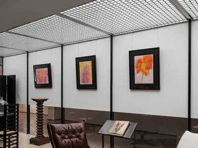 Modern gallery interior with white walls displaying three framed floral watercolor paintings, a brown leather lounge chair with wooden swivel base, a small black side table with a book, a patterned Persian-style rug, and a white sofa in the foreground under a grid skylight.