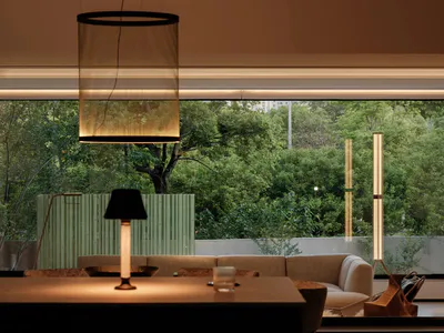 Modern living room interior featuring a large cylindrical sheer fabric pendant light, neutral sofa, table lamp, and expansive windows overlooking dense green trees.