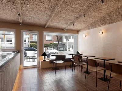 Interior view of a modern cafe with a heavily textured beige plaster ceiling, terracotta tile floor, built-in banquette seating, wooden chairs, and large windows overlooking a street scene.