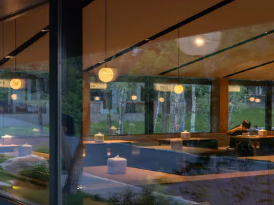 Interior view of a modern Japanese restaurant featuring wooden tables, built-in seating, spherical pendant lights, and a lush forest visible through floor-to-ceiling glass windows.