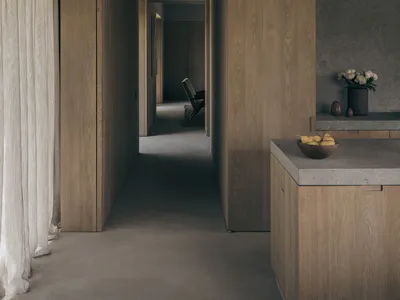 Modern minimalist interior hallway featuring floor-to-ceiling oak wood cabinetry, concrete countertops with a wooden bowl of pears, sheer linen curtain on the left, and a view into a distant living area with a chair.