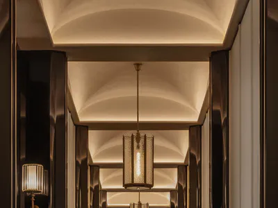 Long narrow luxury hotel hallway with dark wood paneling, arched ceilings with cove lighting, textured cylindrical pendant lanterns, and a colorful abstract painting at the end.