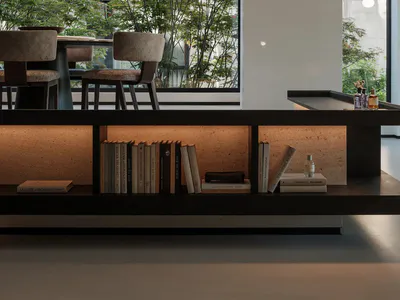 Modern minimalist dining room featuring a large window overlooking green trees, two mesh pendant lights, upholstered dining chairs, and a built-in illuminated travertine bookshelf filled with books in the foreground.
