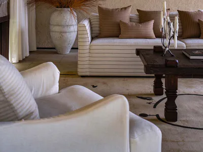 Modern rustic living room interior with white and brown striped sofa, dark wooden coffee table, large white ceramic vase with dried grasses, white armchair in foreground, and textured beige walls under a wood-paneled ceiling.