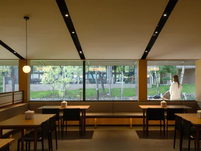 Modern minimalist Japanese restaurant interior featuring light wood tables, black chairs, built-in upholstered banquettes, large windows overlooking a green garden, and spherical pendant lights.