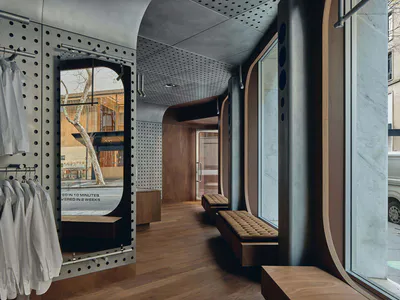 Modern boutique store interior with curved walnut wood walls, built-in upholstered benches, perforated steel clothing racks holding white shirts, and large arched windows overlooking the street.