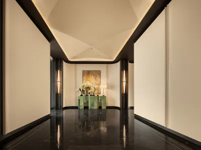 Long elegant hotel hallway with dark marble floor, white walls, dramatic ceiling cove lighting, and a green marble console table with white floral arrangements at the end.