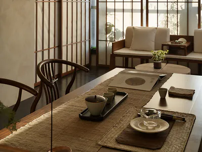 Modern Japanese-style dining room featuring wooden shoji screens, curved wooden dining chairs, a long wooden table with woven placemats, a black pendant light, and a built-in window seat with cushions overlooking bamboo plants.
