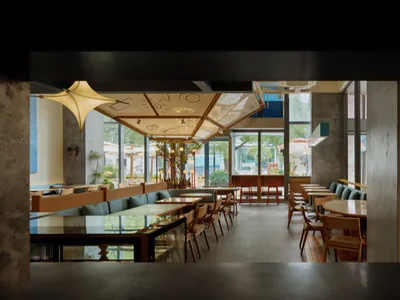 Modern cafe interior viewed from the entrance, showing wooden tables and chairs, teal booth seating, large glass windows, concrete pillars, and a decorative heart-shaped door with the text 'lucky you' on the right side.