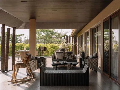Modern tropical covered patio featuring dark woven outdoor sofas, quilted armchairs, a wooden rocking horse, and natural tree-trunk columns overlooking a lush garden.