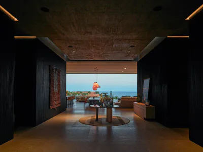 Modern luxury villa interior hallway with dark textured walls, raw concrete ceiling, circular rug, and stunning ocean view in the background.