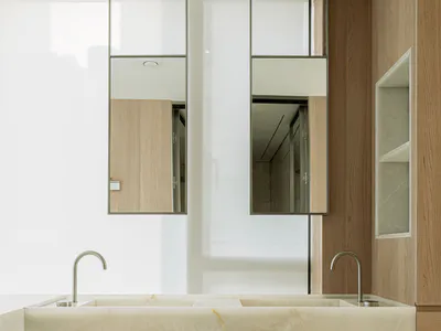 Modern minimalist bathroom with a large white marble double sink vanity supported by a central pedestal, two curved chrome faucets, warm light wood cabinetry, and two tall floating mirrors with metallic frames against a bright white wall.