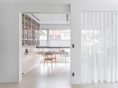 Modern minimalist home office featuring light wood built-in storage, a wooden desk and chair, large windows with roller blinds, and sheer white curtains on the right.