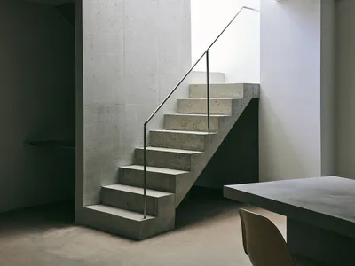Minimalist interior featuring a raw concrete staircase with thin metal handrail and matching concrete dining table.