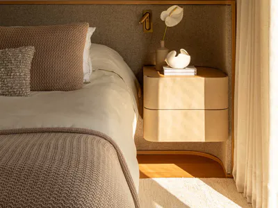 Modern minimalist bedroom interior with a beige upholstered headboard, floating two-drawer walnut nightstand, sculptural brass and alabaster pendant light, and neutral textured bedding.