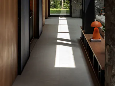 Long modern interior hallway featuring dark wood paneling on the left, a textured stone wall on the right, light concrete floor, wooden ceiling, and a large glass door opening to a green garden with trees. An orange table lamp sits on a wooden console bench in the foreground.