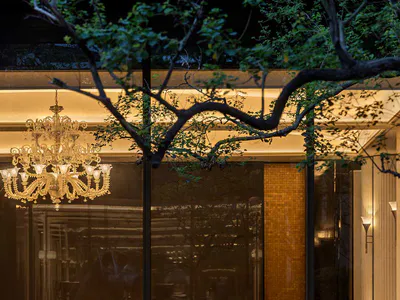 Evening view of a luxurious hotel lobby interior featuring a large ornate crystal chandelier, modern curved sofas, marble and brass accents, and a reflecting pool in the foreground with tree branches framing the scene.