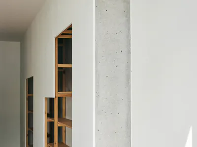 Narrow minimalist hallway featuring built-in oak shelving units recessed into a white wall beside a raw concrete pillar, with light wood flooring.