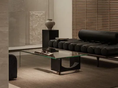 Modern minimalist living room featuring a black leather cylindrical bolster daybed, glass coffee table, concrete walls, and a textured stone sculpture on a black pedestal.