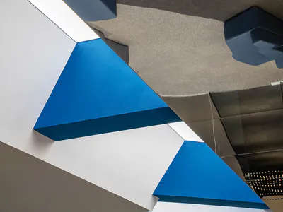 View looking up at a modern geometric ceiling installation made of white, light grey, dark blue, and bright blue felt acoustic panels, with integrated lighting and a small white table and chair visible on the upper level.