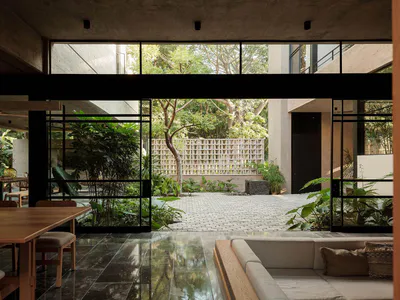 Modern concrete house interior with wooden dining table, light sofa, dark tiled floor, and large glass openings revealing a green courtyard garden with trees and plants.