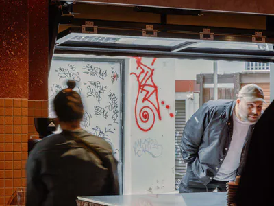 Interior view of a modern cafe showing a textured stone service counter, a barista with hair in a bun facing away, and a bearded man in a cap leaning on the counter. The background features white walls covered in colorful graffiti and red street art, with warm orange lighting and an open pass-through window.
