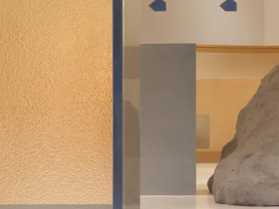 Close-up view of a textured ochre plaster wall next to a blue-framed glass panel, with abstract concrete sculptures and geometric blue wall details in the background.