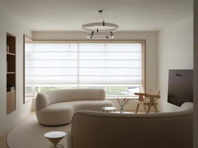 Minimalist living room featuring a large curved boucle sofa, matching armchairs, a circular ring chandelier, and a large window with sheer blinds overlooking greenery.