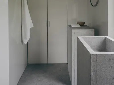 Minimalist bathroom interior with concrete bathtub and vanity, white towel hanging on the wall, round mirror, and tall storage cabinet under soft natural light.