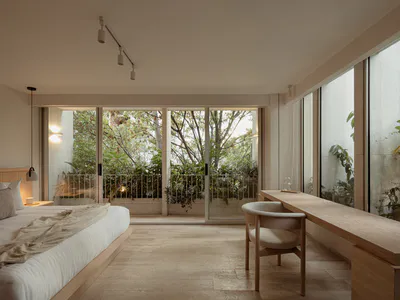 Modern minimalist bedroom featuring a low platform bed with beige linens, a long oak wood desk with a curved upholstered chair, large sliding glass doors revealing a lush green garden, and warm track lighting.