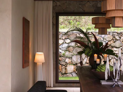Modern rustic dining room featuring a dark wood banquette with upholstered seating, a large wooden dining table, layered wooden pendant lights, a stone wall with large window overlooking a garden, and tropical floral arrangement with candles.