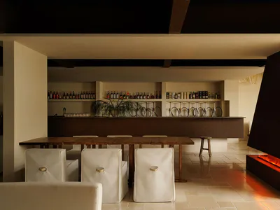 Modern minimalist interior featuring a long wooden dining table with white upholstered chairs, a dark wood bar counter with wine glass racks and liquor shelves, and a large concrete fireplace with glowing fire on the right.