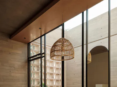 Modern minimalist bathroom featuring a concrete wall and ceiling, wooden rattan pendant light, terrazzo vanity with wooden rattan cabinet, black matte faucet, and large steel windows opening to a courtyard with tropical plants.