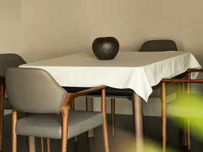 Modern minimalist dining area with walnut wood armchairs upholstered in taupe leather, a rectangular table covered with a white linen tablecloth, and a dark ceramic vase on a dark reflective floor against beige walls.