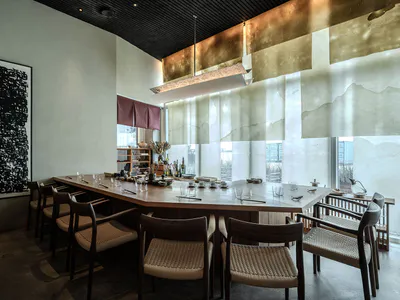 Modern Japanese restaurant interior with a long wooden communal table, dark wood and woven rattan chairs, a large abstract black calligraphy artwork on the left wall, and a striking suspended bronze and white fabric light installation over the table.