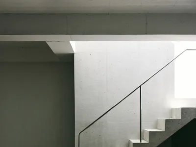 Minimalist brutalist interior with raw concrete walls, a floating concrete staircase with thin metal handrail, and a beige Eames-style chair placed next to a concrete counter.