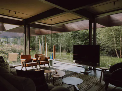 Modern living room interior featuring large glass walls overlooking a forest, orange leather armchairs, wooden ceiling beams, a large flat-screen TV, and a patterned area rug with organic-shaped coffee tables.