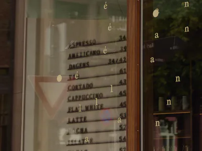 Close-up view of a modern cafe's large glass window featuring gold vinyl lettering spelling coffee-related words, a wooden frame, and reflections of green trees and a neighboring building.