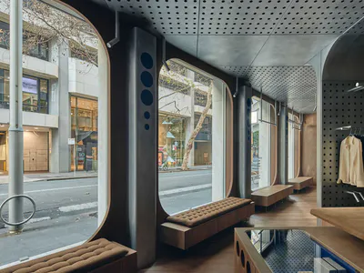 Interior view of a modern clothing boutique with large curved street-facing windows, perforated metal ceiling, wooden benches with brown tufted cushions, and clothing racks against a dark pegboard wall.