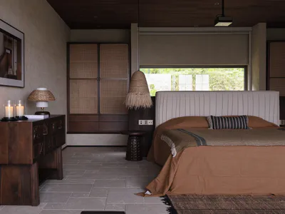 現代臥室內部，大床鋪設赤陶與棕色寢具，懸掛藤編吊燈，深色木質邊櫃上有蠟燭與檯燈，靠牆放置高大的藤編衣櫃，牆面為特殊質感塗料。