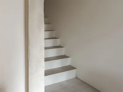 View of a minimalist concrete staircase ascending through a large, hand-finished arched opening in a white plastered wall.