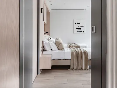 Modern minimalist bedroom viewed through a dark grey doorway, featuring a platform bed with white linens, beige throw blanket, light oak built-in wardrobe and nightstand, and abstract black-and-white wall art.