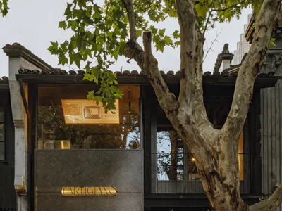 Street view of a modern cafe named Carigiano with large glass windows, concrete upper facade, warm interior lighting, outdoor seating, and a large tree with whitewashed trunk in front of traditional Chinese architecture.
