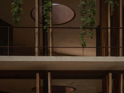 View of a double-height tropical modern living room featuring exposed concrete walls, warm wooden columns, hanging philodendron vines, and large glass doors opening to a lush garden.