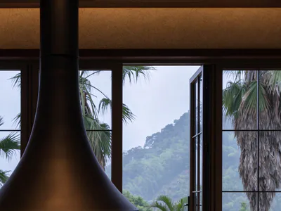 Modern conical steel fireplace with burning logs in a glass enclosure, positioned next to a tan upholstered armchair with striped pillow, opening to a tropical landscape with palm trees, infinity pool, and misty green mountains.