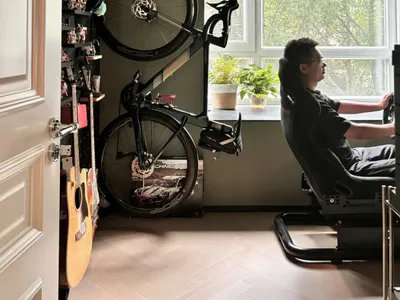 一名男子坐在黑色電競椅上使用電腦，房間內有壁掛公路自行車、木吉他、收藏品和大片採光窗。