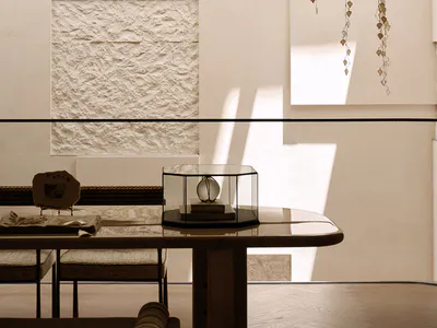 Modern minimalist dining room interior with a large travertine textured wall panel, a dark sculptural pendant lamp, a delicate metal leaf mobile hanging from the ceiling, a marble dining table with a glass display case, and woven dining chairs.